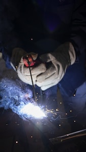 A person wearing protective gloves is using a welding tool, producing bright sparks and smoke. The scene is dimly lit, highlighting the intense glow of the welding process.