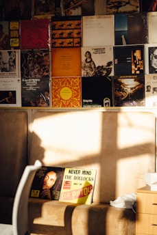 A wall covered with vinyl album covers, featuring a range of artists and styles. In the foreground, two prominent albums are partially illuminated by sunlight, one with a cover of Bob Marley’s album 'Legend' and the other 'Never Mind the Bollocks' by the Sex Pistols. The setting includes a beige seat or couch and a wooden side table, with sunlight casting a shadow across the scene.