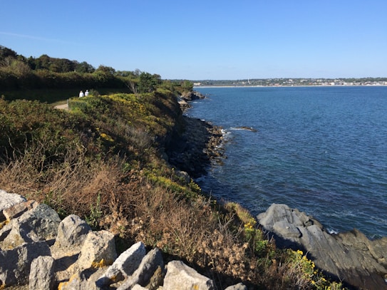 A coastal landscape features a rugged, rocky shoreline with lush greenery and small trees. A narrow path winds through the vegetation, where two people are walking. The blue ocean extends to the horizon, meeting a distant shoreline with a scattering of buildings under a clear blue sky.