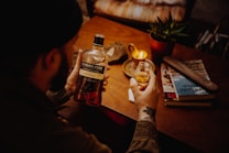 A person is holding a glass of whiskey in one hand and a bottle of 'Highland Park' whiskey in the other. The setting includes a cozy wooden table with a lit candle, a potted plant, and a stack of books. The scene is warmly lit, creating an inviting atmosphere.