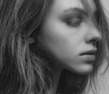 A close-up black and white portrait of a person looking to the side, with hair falling naturally around the face. The lighting highlights the contours and textures of the skin and hair, creating a soft and detailed visual effect.