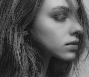 A close-up black and white portrait of a person looking to the side, with hair falling naturally around the face. The lighting highlights the contours and textures of the skin and hair, creating a soft and detailed visual effect.
