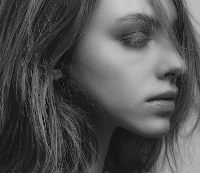 A close-up black and white portrait of a person looking to the side, with hair falling naturally around the face. The lighting highlights the contours and textures of the skin and hair, creating a soft and detailed visual effect.