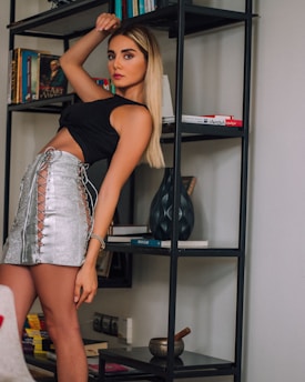 A woman with long blonde hair is leaning against a black metal bookshelf filled with books and decorative items. She is wearing a black crop top and a silver skirt with lace-up sides. The background includes several other books and a decorative vase.
