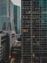 An urban scene featuring multiple modern high-rise buildings with glass exteriors. The buildings appear to be offices, with lights turned on in some windows suggesting occupancy or activity. There is a noticeable contrast between the different architectural designs of the buildings including straight lines and geometric patterns.