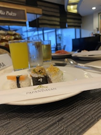 A close-up of a plate featuring assorted sushi rolls on a dining table. A pair of neatly wrapped chopsticks with 'The Papandayan' branding is placed beside the plate. In the background, there are glasses filled with iced beverages. The overall setting suggests a dining establishment with seating and ambient lighting.