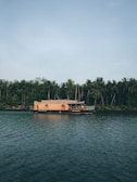 A traditional houseboat floats on a calm river with lush greenery and dense forest trees in the background, creating a peaceful and serene setting.