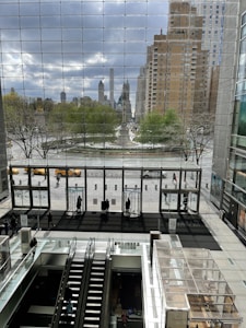 The image features an indoor shopping mall with large glass windows providing a view of the cityscape outside. Beyond the glass windows, there is a park with green trees and various buildings in the background, some of which are tall skyscrapers. The interior includes an escalator leading down to a lower floor and several people walking or standing near the entrance doors. Yellow taxis and a few pedestrians are visible outside.