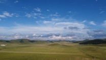 Expansive open landscape with gentle rolling hills covered in green grass. A large expanse of blue sky with scattered white clouds stretches across the horizon. Sparse trees are visible along the hills, giving a sense of wide, natural space.