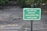 A green sign with white text stating 'DISEASED TREE REMOVED REPLACEMENT TREE TO BE PLANTED' is placed in a dirt area with sparse grass and some scattered debris. There are trees and greenery in the blurred background.