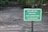 A green sign with white text stating 'DISEASED TREE REMOVED REPLACEMENT TREE TO BE PLANTED' is placed in a dirt area with sparse grass and some scattered debris. There are trees and greenery in the blurred background.