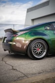 Modified sports car with a shiny, color-changing paint job ranging from green to brown, parked on an asphalt lot near a chain-link fence. The back wheel features custom rims with red detailing, and a small character decal is visible on the side window.