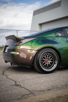 Modified sports car with a shiny, color-changing paint job ranging from green to brown, parked on an asphalt lot near a chain-link fence. The back wheel features custom rims with red detailing, and a small character decal is visible on the side window.