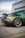 Modified sports car with a shiny, color-changing paint job ranging from green to brown, parked on an asphalt lot near a chain-link fence. The back wheel features custom rims with red detailing, and a small character decal is visible on the side window.