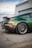 Modified sports car with a shiny, color-changing paint job ranging from green to brown, parked on an asphalt lot near a chain-link fence. The back wheel features custom rims with red detailing, and a small character decal is visible on the side window.