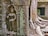 Intricately carved stone relief featuring a figure in traditional attire is set against the backdrop of ancient ruins. Large tree roots envelop the structure, showcasing the interplay between nature and architecture.