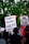 A group of people at a protest hold signs advocating for justice and criticizing law enforcement practices. The visible signs have messages calling for a people's verdict and addressing racial issues. The background includes lush green trees and a traffic light.