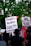 A group of people at a protest hold signs advocating for justice and criticizing law enforcement practices. The visible signs have messages calling for a people's verdict and addressing racial issues. The background includes lush green trees and a traffic light.