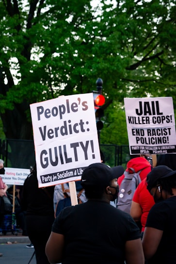 A group of people at a protest hold signs advocating for justice and criticizing law enforcement practices. The visible signs have messages calling for a people's verdict and addressing racial issues. The background includes lush green trees and a traffic light.