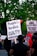 A group of people at a protest hold signs advocating for justice and criticizing law enforcement practices. The visible signs have messages calling for a people's verdict and addressing racial issues. The background includes lush green trees and a traffic light.