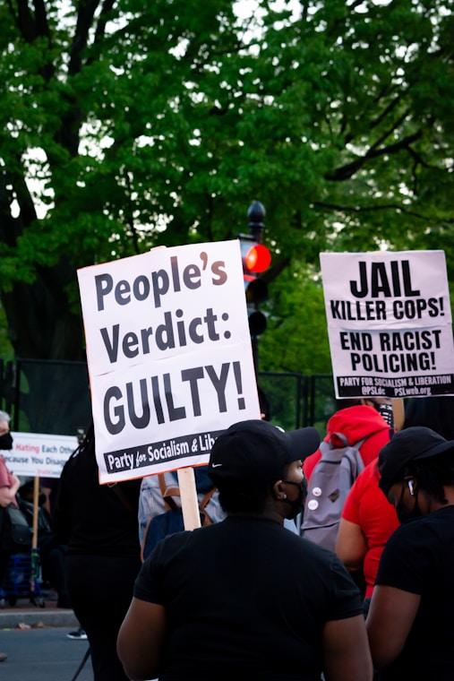 A group of people at a protest hold signs advocating for justice and criticizing law enforcement practices. The visible signs have messages calling for a people's verdict and addressing racial issues. The background includes lush green trees and a traffic light.