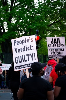 A group of people at a protest hold signs advocating for justice and criticizing law enforcement practices. The visible signs have messages calling for a people's verdict and addressing racial issues. The background includes lush green trees and a traffic light.