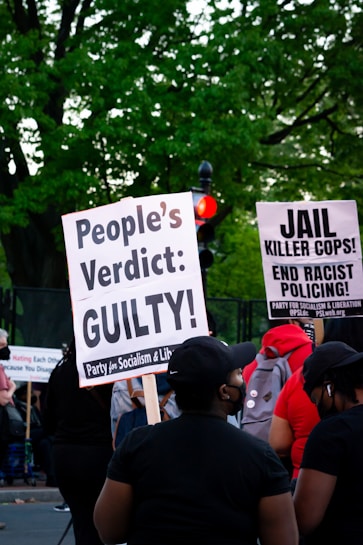 A group of people at a protest hold signs advocating for justice and criticizing law enforcement practices. The visible signs have messages calling for a people's verdict and addressing racial issues. The background includes lush green trees and a traffic light.