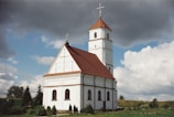 A white church with a red-tiled roof and a tall tower topped by a cross is set against a picturesque rural landscape. The building features arched windows and simple yet elegant architectural details. An overcast sky with both dark and fluffy clouds creates a dramatic backdrop. Surrounding the church, lush green grass and small shrubs add to the serene environment.