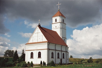 A white church with a red-tiled roof and a tall tower topped by a cross is set against a picturesque rural landscape. The building features arched windows and simple yet elegant architectural details. An overcast sky with both dark and fluffy clouds creates a dramatic backdrop. Surrounding the church, lush green grass and small shrubs add to the serene environment.
