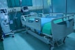 A modern hospital room featuring an adjustable hospital bed with side rails, a blue sheet, and medical devices such as monitors and an infusion pump nearby. The room is tiled, with a curtain in the background and medical equipment along the walls.