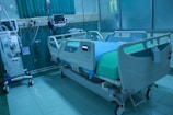 A modern hospital room featuring an adjustable hospital bed with side rails, a blue sheet, and medical devices such as monitors and an infusion pump nearby. The room is tiled, with a curtain in the background and medical equipment along the walls.