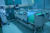 A modern hospital room featuring an adjustable hospital bed with side rails, a blue sheet, and medical devices such as monitors and an infusion pump nearby. The room is tiled, with a curtain in the background and medical equipment along the walls.