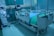 A modern hospital room featuring an adjustable hospital bed with side rails, a blue sheet, and medical devices such as monitors and an infusion pump nearby. The room is tiled, with a curtain in the background and medical equipment along the walls.