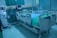 A modern hospital room featuring an adjustable hospital bed with side rails, a blue sheet, and medical devices such as monitors and an infusion pump nearby. The room is tiled, with a curtain in the background and medical equipment along the walls.