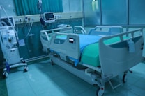 A modern hospital room featuring an adjustable hospital bed with side rails, a blue sheet, and medical devices such as monitors and an infusion pump nearby. The room is tiled, with a curtain in the background and medical equipment along the walls.