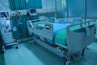 A modern hospital room featuring an adjustable hospital bed with side rails, a blue sheet, and medical devices such as monitors and an infusion pump nearby. The room is tiled, with a curtain in the background and medical equipment along the walls.