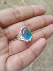A hand holding a silver ring with a large, oval-shaped stone that has an iridescent blue-green hue.