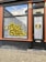 The image shows the exterior of a building with a large window and a door. The window displays gold letter balloons spelling out 'JESUS CHRIST LOVE YOU.' There are also two posters with images and text next to the balloon letters. The door has a curtain behind it and a small mail slot at the bottom. The building has red-orange columns on either side of the window and door, with a tiled mosaic near the top.