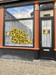 The image shows the exterior of a building with a large window and a door. The window displays gold letter balloons spelling out 'JESUS CHRIST LOVE YOU.' There are also two posters with images and text next to the balloon letters. The door has a curtain behind it and a small mail slot at the bottom. The building has red-orange columns on either side of the window and door, with a tiled mosaic near the top.