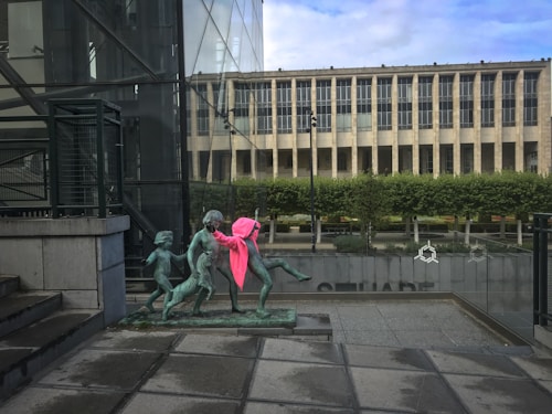 A modern public art sculpture of children is located outdoors near a glass building. One child is adorned with a bright pink hoodie. In the background, a formal building with numerous tall windows stands, surrounded by well-maintained greenery.