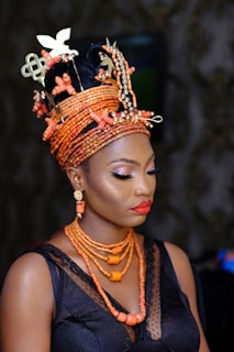 A woman is wearing an elaborate headdress made of coral beads with ornate metallic designs and bright makeup. Her attire includes a black lace top and coral bead necklaces, coordinated with matching earrings. Her expression is serene and contemplative.