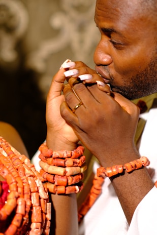 A person wearing traditional ornaments, including multiple coral bead necklaces and bracelets, is holding hands intimately with another individual. The scene conveys a sense of affection and cultural richness, as both individuals are engaged in a tender gesture.