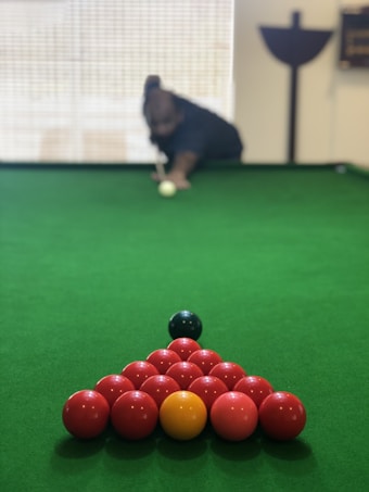 A close-up view of a snooker or pool table, with a triangle of red balls and a single yellow ball set up at the center. A cue ball is positioned in the foreground, and a person is seen in the background poised to take a shot.