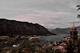 A picturesque coastal town is nestled between large, rugged mountains and a serene body of water. The town, with its cluster of buildings and a prominent church tower, sprawls along the shoreline. A cruise ship glides through the peaceful waters, adding a sense of travel and exploration. Lush greenery frames the scene, suggesting a natural, tranquil environment.