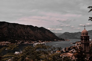 A picturesque coastal town is nestled between large, rugged mountains and a serene body of water. The town, with its cluster of buildings and a prominent church tower, sprawls along the shoreline. A cruise ship glides through the peaceful waters, adding a sense of travel and exploration. Lush greenery frames the scene, suggesting a natural, tranquil environment.
