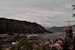 A picturesque coastal town is nestled between large, rugged mountains and a serene body of water. The town, with its cluster of buildings and a prominent church tower, sprawls along the shoreline. A cruise ship glides through the peaceful waters, adding a sense of travel and exploration. Lush greenery frames the scene, suggesting a natural, tranquil environment.