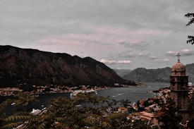 A picturesque coastal town is nestled between large, rugged mountains and a serene body of water. The town, with its cluster of buildings and a prominent church tower, sprawls along the shoreline. A cruise ship glides through the peaceful waters, adding a sense of travel and exploration. Lush greenery frames the scene, suggesting a natural, tranquil environment.