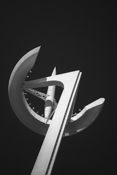 A modern, abstract architectural structure with a tall vertical element and curved, wing-like extensions against a dark background. The design incorporates geometric shapes and sharp angles, creating a sense of futuristic style.