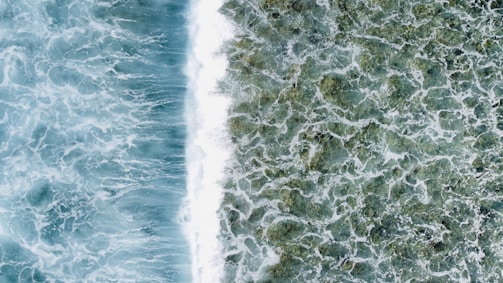 Two distinct sections of ocean waves from a top-down perspective, with the left side showing deeper blue, turbulent water and the right side displaying lighter, foamy waves over shallower green water.