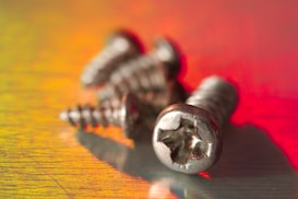 Several metallic screws lie scattered on a surface that reflects warm hues of red, orange, and yellow, creating a vivid and vibrant backdrop. The foreground is sharply in focus, displaying the intricate details of the screw heads.